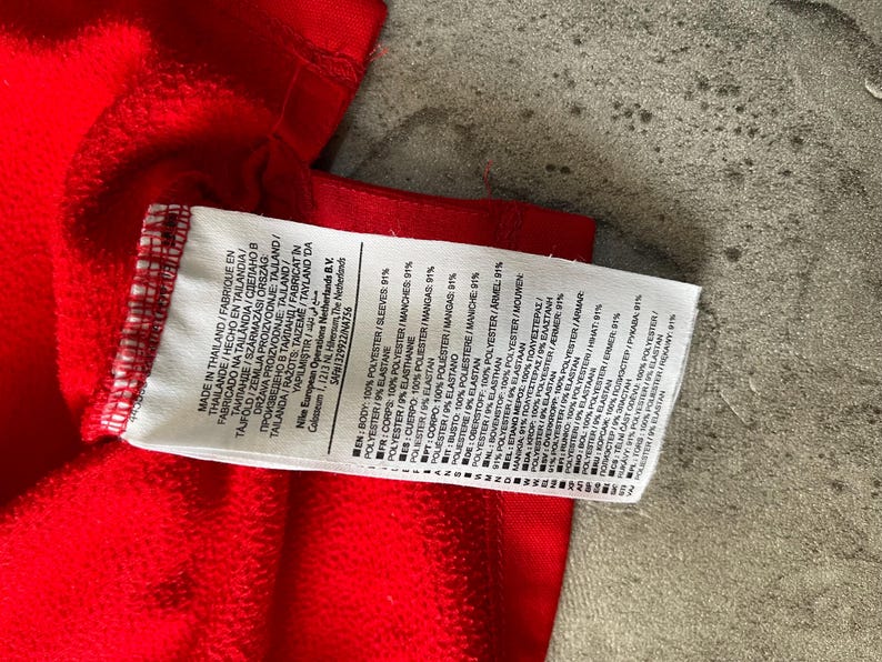 May include: A close-up of a red garment with a white tag attached. The tag displays text in multiple languages, including English, French, and Spanish, detailing the fabric composition as 100% polyester and 9% elastane.