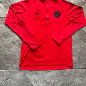 May include: A red long-sleeved athletic shirt with a quarter-zip collar. Features a black Jordan logo and a Paris Saint-Germain emblem on the chest. The shirt has black stripes on the sleeves.