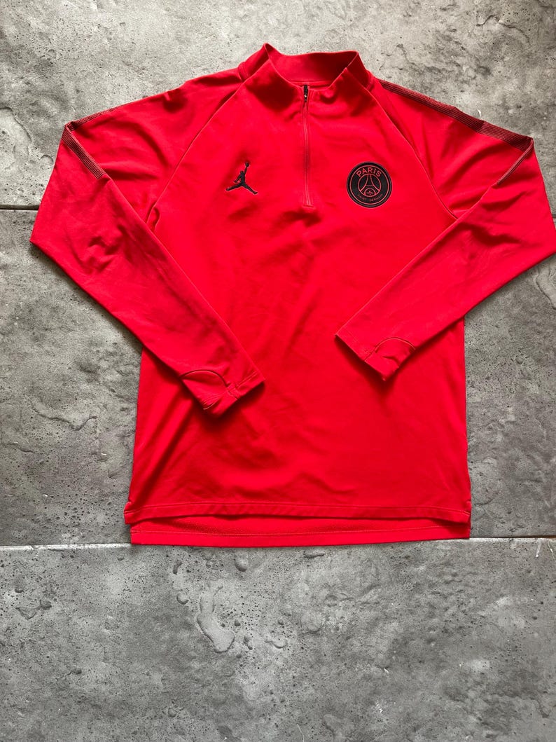 May include: A red long-sleeved athletic shirt with a quarter-zip collar. Features a black Jordan logo and a Paris Saint-Germain emblem on the chest. The shirt has black stripes on the sleeves.