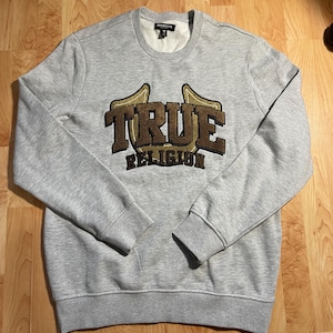 Puede incluir: Una sudadera gris claro con el texto "TRUE RELIGION" en un diseño marrón y dorado. La sudadera tiene cuello redondo y puños y dobladillo acanalados. El nombre de la marca se muestra en la parte delantera.
