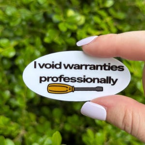 May include: White oval sticker with the text "I void warranties professionally" in black. A yellow and black screwdriver graphic is below the text. The sticker is held by a person with light purple nail polish.