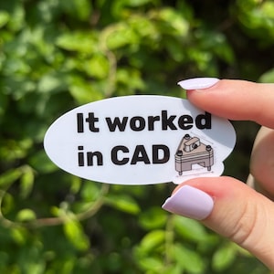 May include: White oval sticker with the text "It worked in CAD" in black, and a small illustration of a mechanical component. The sticker is held against a background of green foliage.