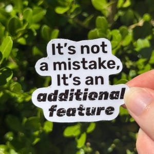 May include: A white sticker with black text that reads "It's not a mistake, It's an additional feature." The sticker is held against a backdrop of green foliage.