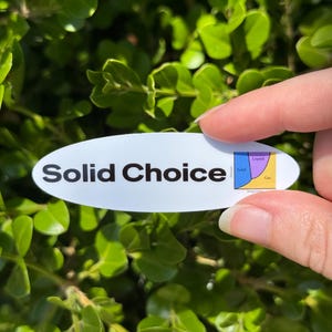 May include: An oval white sticker with the words "Solid Choice" in black text. A small diagram showing phases of matter is on the right. The sticker is held in front of a background of green leaves.