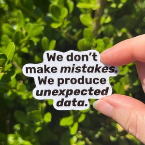 May include: A white sticker with black text that reads "We don't make mistakes. We produce unexpected data." The sticker is held in front of a background of green leaves.