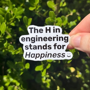 May include: White sticker with black text that reads "The H in engineering stands for Happiness :) ". The sticker is held in front of a background of green leaves.