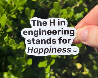 May include: White sticker with black text that reads "The H in engineering stands for Happiness :) ". The sticker is held in front of a background of green leaves.