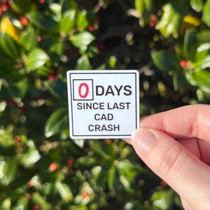 Peut inclure: Un autocollant carré blanc avec des bordures noires et le texte "0 JOURS DEPUIS LE DERNIER PLANTAGE CAO". Le chiffre "0" est en rouge. L'autocollant est tenu devant un fond de feuilles vertes.