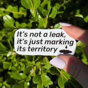 May include: A white sticker with the text "It's not a leak, it's just marking its territory" in black. A small illustration of a water droplet and a puddle is on the right. The sticker is held against a background of green leaves.
