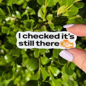 May include: A white sticker with black text that reads "I checked it's still there." An orange "CHECK ENGINE" light graphic is on the right. The sticker is held in front of a green leafy background.