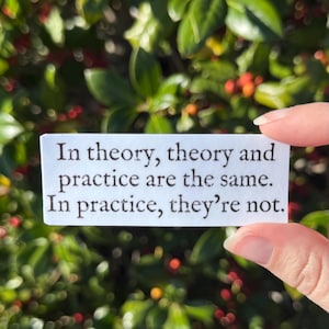 May include: A white rectangular sticker with the text "In theory, theory and practice are the same. In practice, they're not." The sticker is held against a background of green leaves and red berries.