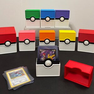 May include: A collection of rectangular trading card storage boxes designed like Poké Balls in various colors including red, green, blue, purple, pink, orange, and yellow. Each box features a white base and a central circular button detail.