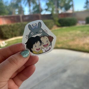 Puede incluir: Una pegatina transparente con una imagen de dibujos animados de Totoro y dos ni&ntilde;os. Totoro, un gran esp&iacute;ritu del bosque gris, sonr&iacute;e ampliamente, y los ni&ntilde;os tambi&eacute;n sonr&iacute;en. La pegatina se sostiene contra un fondo borroso al aire libre.