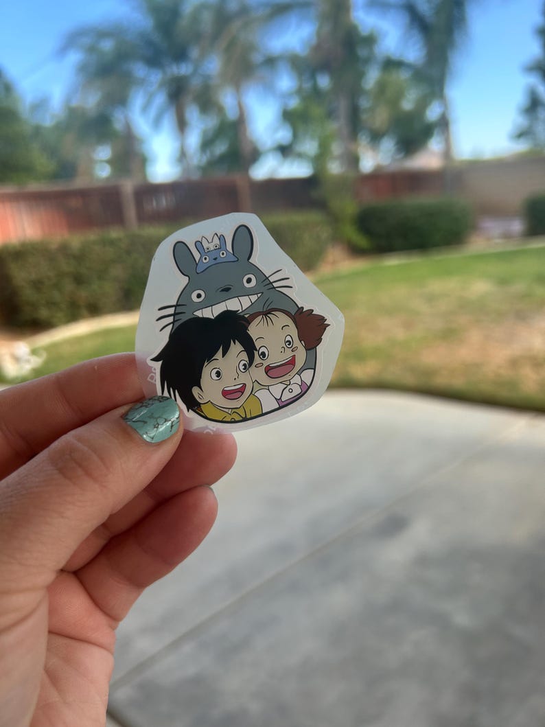 Puede incluir: Una pegatina transparente con una imagen de dibujos animados de Totoro y dos ni&ntilde;os. Totoro, un gran esp&iacute;ritu del bosque gris, sonr&iacute;e ampliamente, y los ni&ntilde;os tambi&eacute;n sonr&iacute;en. La pegatina se sostiene contra un fondo borroso al aire libre.