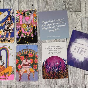 May include: A set of tarot cards with colorful illustrations and text. The cards feature a rainbow, a lion, a person sitting on a throne, and a person holding a sword. The cards are on a wooden surface. The text on the cards includes "Strength", "IX", "Practice the Pause", and "I can decide today to recalibrate my energy and commit to love and joy."