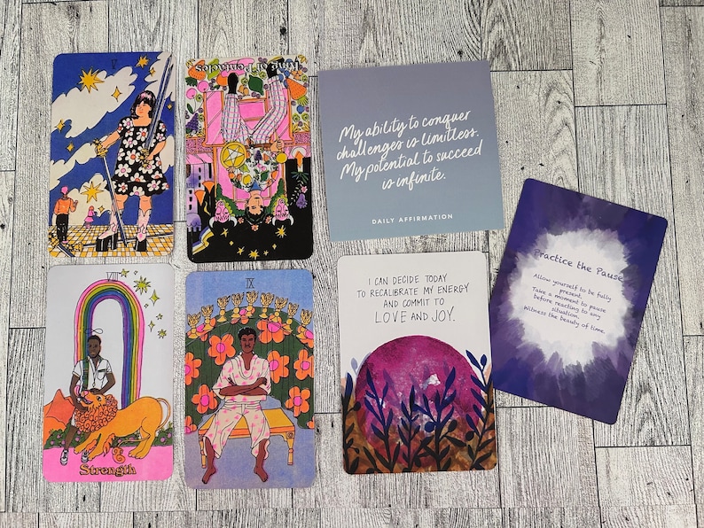 May include: A set of tarot cards with colorful illustrations and text. The cards feature a rainbow, a lion, a person sitting on a throne, and a person holding a sword. The cards are on a wooden surface. The text on the cards includes "Strength", "IX", "Practice the Pause", and "I can decide today to recalibrate my energy and commit to love and joy."