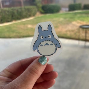 Puede incluir: Una pegatina azul y blanca con un personaje de dibujos animados con ojos grandes y una barriga esponjosa. La pegatina tiene un borde transparente y se sostiene contra un fondo borroso al aire libre. El personaje tiene una expresi&oacute;n amigable.