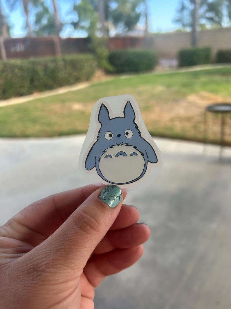 Puede incluir: Una pegatina azul y blanca con un personaje de dibujos animados con ojos grandes y una barriga esponjosa. La pegatina tiene un borde transparente y se sostiene contra un fondo borroso al aire libre. El personaje tiene una expresi&oacute;n amigable.