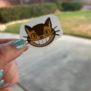 Puede incluir: Una pegatina transparente con la cara de un gato de dibujos animados con una gran sonrisa, pelaje marr&oacute;n y ojos amarillos. La pegatina se sostiene contra un fondo borroso al aire libre. El dise&ntilde;o del gato recuerda a un personaje animado popular.