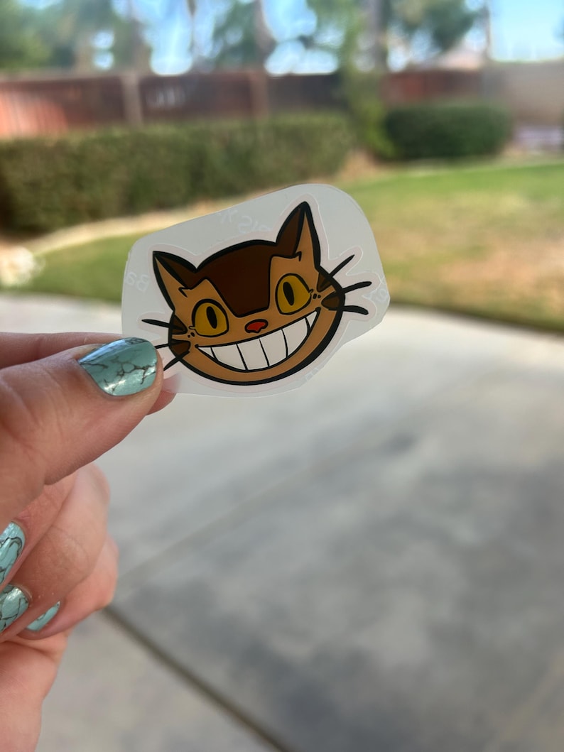 Puede incluir: Una pegatina transparente con la cara de un gato de dibujos animados con una gran sonrisa, pelaje marr&oacute;n y ojos amarillos. La pegatina se sostiene contra un fondo borroso al aire libre. El dise&ntilde;o del gato recuerda a un personaje animado popular.