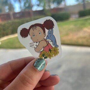 Puede incluir: Una pegatina transparente con una ni&ntilde;a de dibujos animados con coletas marrones, un vestido rosa y una criatura azul. La ni&ntilde;a est&aacute; rodeada de flores amarillas. La pegatina est&aacute; sujeta por una mano con esmalte de u&ntilde;as turquesa.