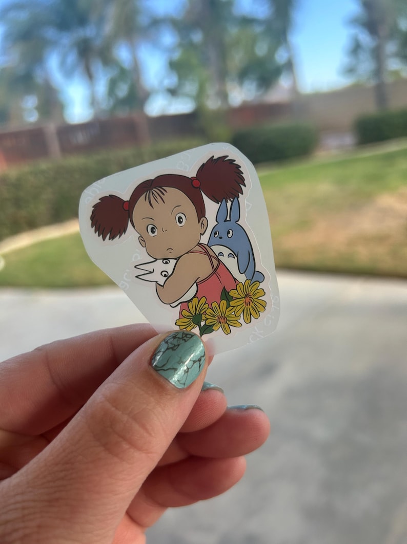 Puede incluir: Una pegatina transparente con una ni&ntilde;a de dibujos animados con coletas marrones, un vestido rosa y una criatura azul. La ni&ntilde;a est&aacute; rodeada de flores amarillas. La pegatina est&aacute; sujeta por una mano con esmalte de u&ntilde;as turquesa.
