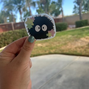 Puede incluir: Una pegatina negra con un sprite de holl&iacute;n de dibujos animados con ojos blancos grandes y acentos coloridos en forma de estrella. La pegatina tiene un borde blanco y se sostiene sobre un fondo de hierba verde y palmeras.
