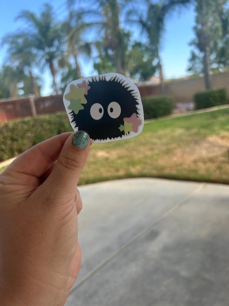 Puede incluir: Una pegatina negra con un sprite de holl&iacute;n de dibujos animados con ojos blancos grandes y acentos coloridos en forma de estrella. La pegatina tiene un borde blanco y se sostiene sobre un fondo de hierba verde y palmeras.