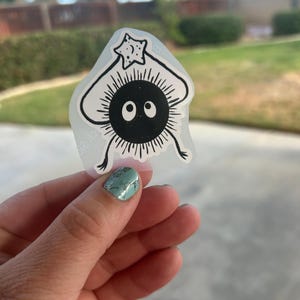 Puede incluir: Una pegatina en blanco y negro que representa un personaje de dibujos animados con un cuerpo redondo, ojos grandes y un objeto en forma de estrella sobre su cabeza. La pegatina tiene un fondo transparente.