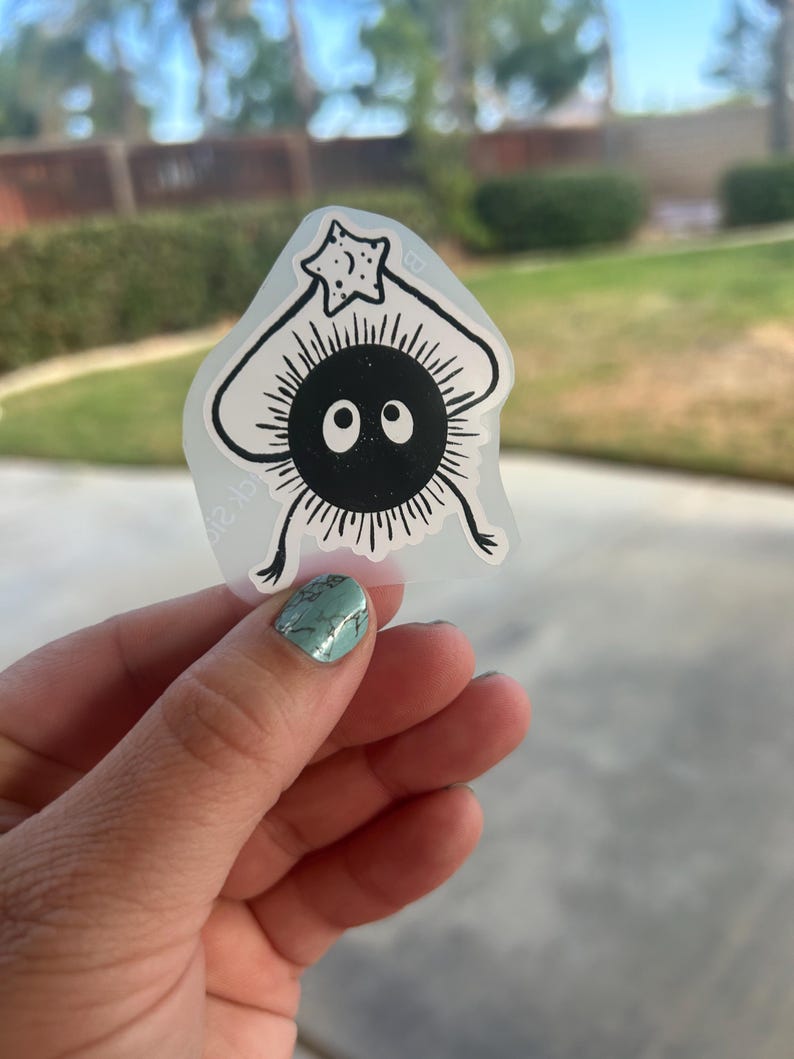 Puede incluir: Una pegatina en blanco y negro que representa un personaje de dibujos animados con un cuerpo redondo, ojos grandes y un objeto en forma de estrella sobre su cabeza. La pegatina tiene un fondo transparente.