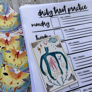 K&ouml;nnte beinhalten: Ein t&auml;glicher Tarot-Praxisplaner mit einer handgezeichneten Tarotkarte, die eine blaue Figur mit einer rosa Figur im Inneren zeigt, umgeben von Kreisen und einer Pflanze mit Bl&auml;ttern und zwei W&ouml;lfen. Der Planer hat ein schwarz-wei&szlig;es Design mit den Worten "t&auml;gliche Tarot-Praxis", "Montag" und "Dienstag" in Schwarz.