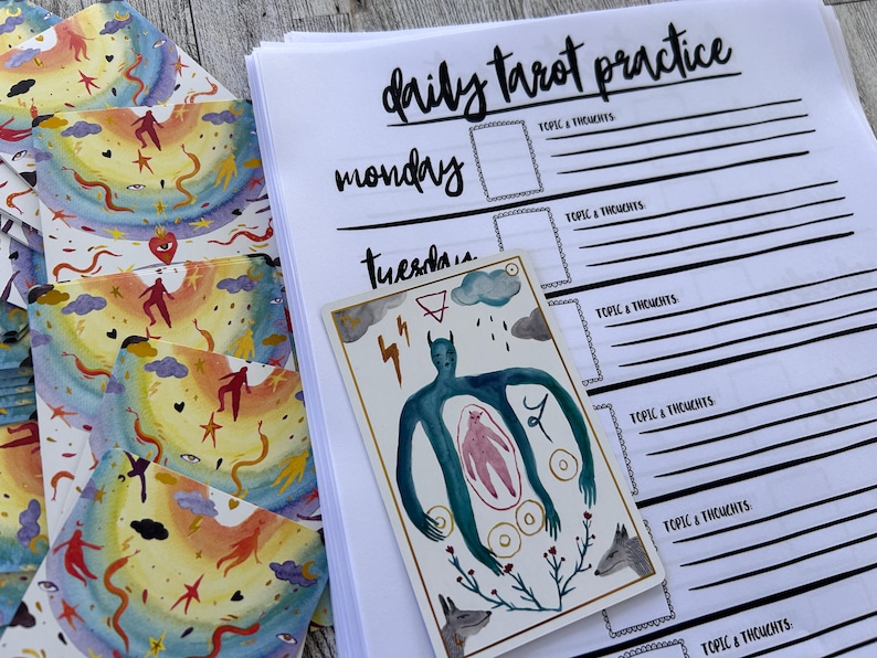 K&ouml;nnte beinhalten: Ein t&auml;glicher Tarot-Praxisplaner mit einer handgezeichneten Tarotkarte, die eine blaue Figur mit einer rosa Figur im Inneren zeigt, umgeben von Kreisen und einer Pflanze mit Bl&auml;ttern und zwei W&ouml;lfen. Der Planer hat ein schwarz-wei&szlig;es Design mit den Worten "t&auml;gliche Tarot-Praxis", "Montag" und "Dienstag" in Schwarz.