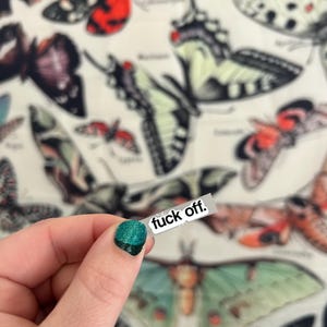 May include: A hand holding a small sticker with the words "fuck off." The background features a fabric with various butterfly illustrations in shades of red, green, black, and white. The person's fingernail is painted with green glitter nail polish.
