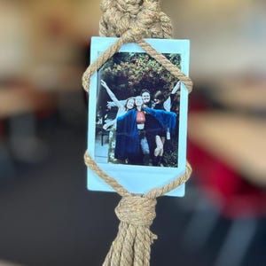 May include: A macrame photo holder with a light blue backing holding a photo of four people. The macrame is made of a tan, sparkly rope, with a knotted design and a tassel at the bottom. The photo shows people posing outdoors.