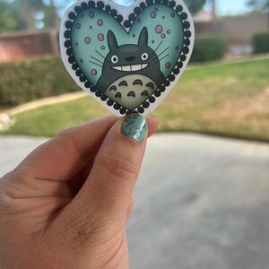Puede incluir: Una pegatina en forma de coraz&oacute;n con un personaje de dibujos animados con un cuerpo gris, dientes blancos y una expresi&oacute;n sonriente. El fondo es azul claro con lunares rosas y el borde es negro con puntos blancos.