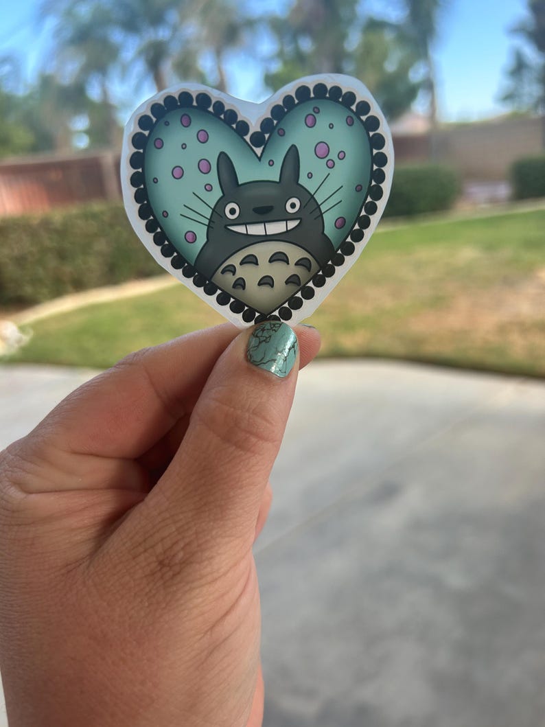 Puede incluir: Una pegatina en forma de coraz&oacute;n con un personaje de dibujos animados con un cuerpo gris, dientes blancos y una expresi&oacute;n sonriente. El fondo es azul claro con lunares rosas y el borde es negro con puntos blancos.