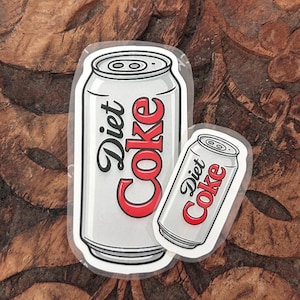Può includere: Due adesivi in vinile fustellati che raffigurano una lattina di bibita argentata con la scritta Diet Coke in lettere nere e rosse. L'adesivo più grande è posizionato verticalmente, mentre quello più piccolo è inclinato sulla destra.