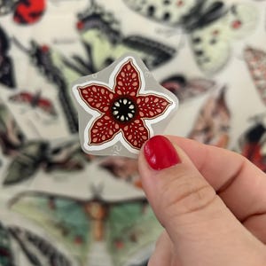 May include: A sticker featuring a red and black Demogorgon flower design with a white outline. The sticker is held in front of a background of various butterfly and moth illustrations.