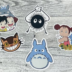 Puede incluir: Una colecci&oacute;n de pegatinas coloridas con personajes de la pel&iacute;cula de animaci&oacute;n "Mi vecino Totoro". Las pegatinas incluyen a Totoro, el autob&uacute;s gato y otros personajes ic&oacute;nicos. Las pegatinas est&aacute;n sobre un fondo de vetas de madera clara.