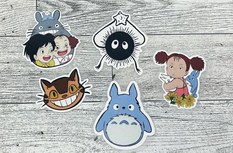 Puede incluir: Una colecci&oacute;n de pegatinas coloridas con personajes de la pel&iacute;cula de animaci&oacute;n "Mi vecino Totoro". Las pegatinas incluyen a Totoro, el autob&uacute;s gato y otros personajes ic&oacute;nicos. Las pegatinas est&aacute;n sobre un fondo de vetas de madera clara.