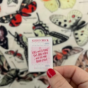 Op de afbeelding: Een kleine witte bon met de tekst "CALL ME A GOOD GIRL AND TAKE ME TO THE BOOKSTORE" wordt vastgehouden voor een achtergrond van vlinderillustraties. De bon heeft een roze rasterpatroon en het nummer 025619.