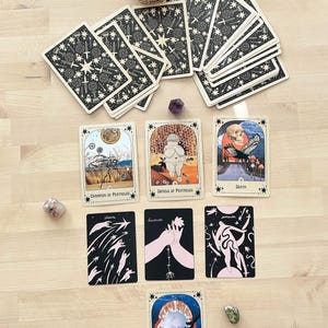 May include: Tarot card spread with 10 cards laid out on a wooden surface. The cards include: Champion of Pentacles, Circle of Pentacles, Death, Stars, Lovers, Surrender, Circle of Cups, and a deck of cards with a star and sword design.