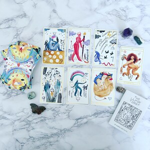 May include: A spread of tarot cards on a white marble surface. The cards are from The Luer Tarot deck and feature colorful illustrations of people, animals, and symbols. The cards are arranged in a circle with a book titled "The Luer Tarot" in the lower right corner.