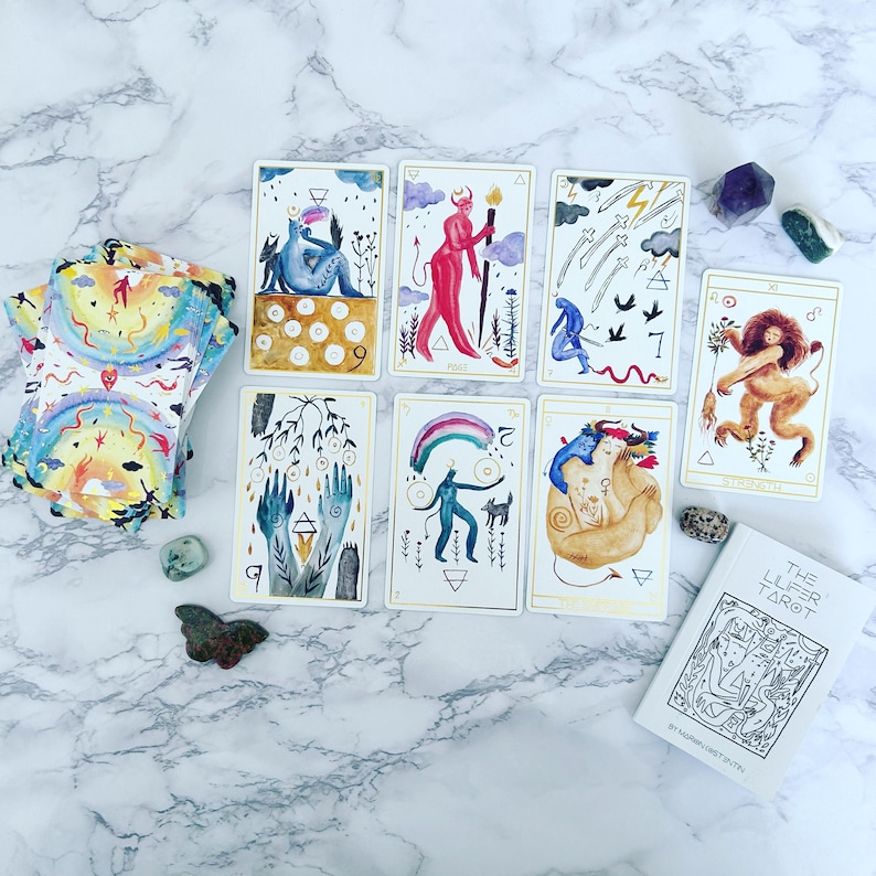 May include: A spread of tarot cards on a white marble surface. The cards are from The Luer Tarot deck and feature colorful illustrations of people, animals, and symbols. The cards are arranged in a circle with a book titled "The Luer Tarot" in the lower right corner.