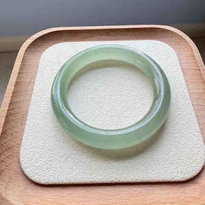 May include: A translucent, pale green jade bangle bracelet. The smooth, polished bracelet is circular and sits on a cream-colored, rectangular surface within a wooden tray. The jade has a subtle, natural color.