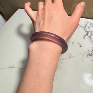 May include: A translucent, lavender-colored bangle bracelet. The smooth, rounded bracelet is a solid color and appears to be made of stone or glass. The bracelet is worn on a wrist.