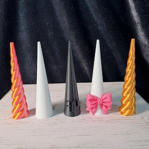 May include: Five unicorn horn-shaped objects in different colours and designs. The horns include a pink and orange swirled horn, a white horn, a black horn, a white horn with a pink bow, and a gold swirled horn.