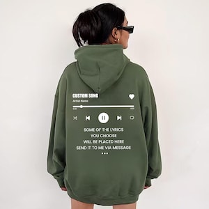May include: An olive green hooded sweatshirt featuring a white graphic on the back that mimics a music player interface. The design includes the text CUSTOM SONG, Artist Name, and the lyrics SOME OF THE LYRICS YOU CHOOSE WILL BE PLACED HERE.