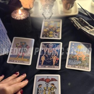 May include: A collection of tarot cards, including "Page of Cups," "The Devil," and "The Star," arranged on a dark surface. A lit black candle and a hand are also visible, suggesting a mystical or spiritual theme.