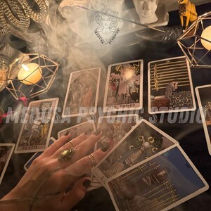 May include: A mystical scene featuring tarot cards, candles, and decorative objects. The cards are spread out, with the hand of a person visible, adorned with rings. The image has a dark, atmospheric aesthetic, with the text "MEDUSA PSYCHIC STUDIO" overlaid.